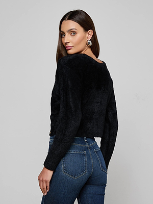 A woman with long brown hair models the L'AGENCE Antonella Crossover Sweater in black with blue jeans and large hoop earrings, standing against a plain gray background and gazing over her shoulder.