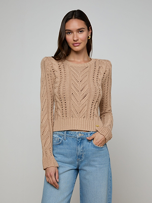 A woman with long brown hair models the Mio Cabled Pullover—a beige cotton-blend knit crewneck sweater with a chevron pattern—and light blue jeans against a plain light gray background, looking at the camera with a slight smile.