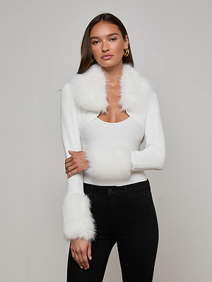 A woman with long brown hair channels old Hollywood glamour in the L'AGENCE Haru Faux Fur Shrug, featuring an ivory hue with removable faux fur collar and cuffs, styled with black pants and gold hoop earrings against a light gray background.