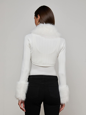 A woman faces away, modeling the L’AGENCE Haru Faux Fur Shrug—a fitted ivory jacket with removable faux fur trim—paired with high-waisted black pants for an Old Hollywood look against a plain, light background.