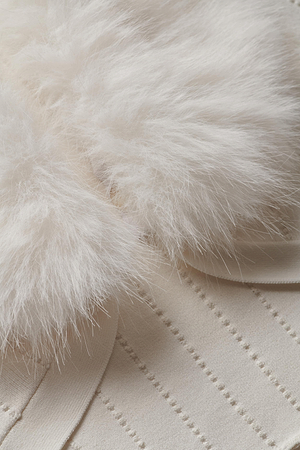 Close-up of two ivory faux fur pom-poms, removable from the Haru Faux Fur Shrug by L'AGENCE, resting on its textured cream fabric with dotted stitching.
