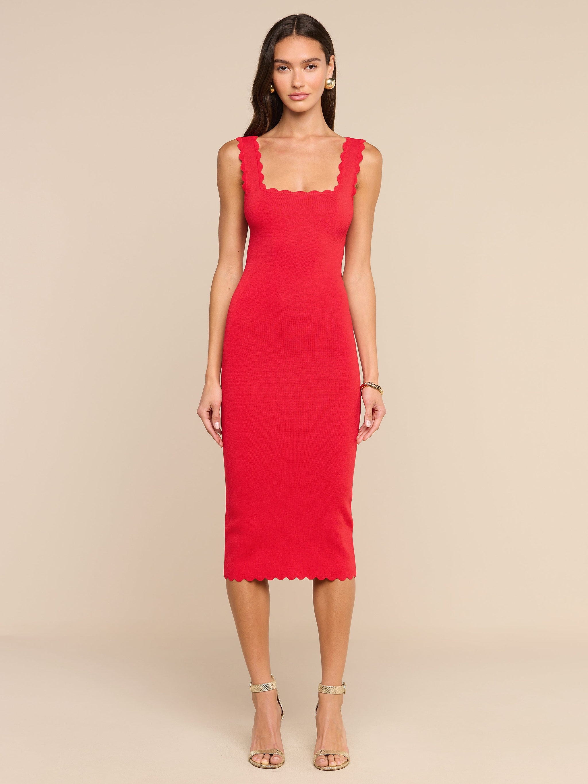 Una donna è in posa su uno sfondo beige e indossa l’abito in maglia Romona: un miniabito senza maniche, dalla linea aderente, di colore rosso rossetto, caratterizzato da scollo quadrato smerlato e orlo smerlato. Ha i capelli lunghi e abbina il look a sandali beige con tacco alto e cinturini e a un braccialetto.