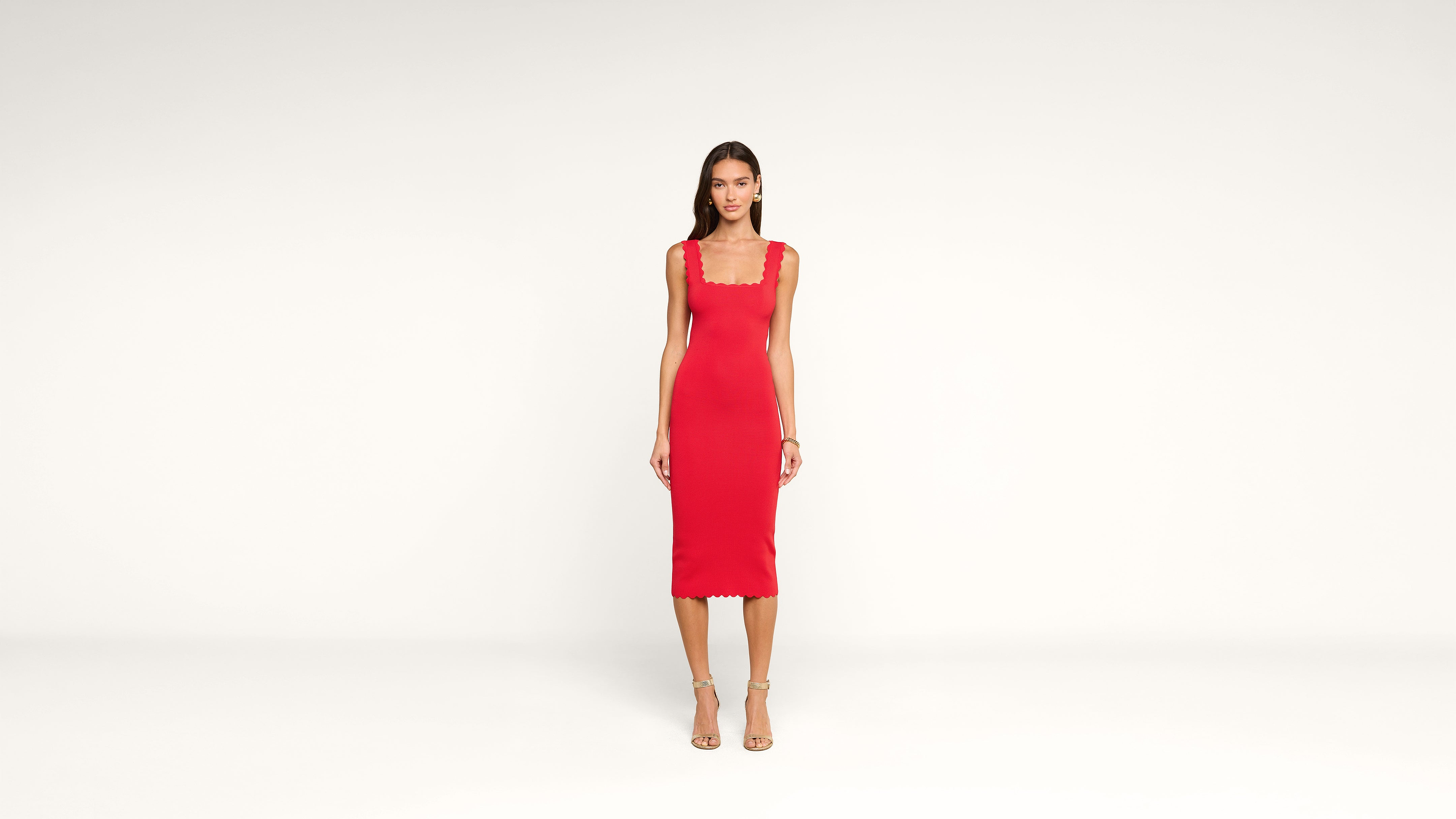 A woman stands against a plain white background wearing a fitted, sleeveless red dress with a square neckline and scalloped edges, paired with strappy high-heeled sandals.