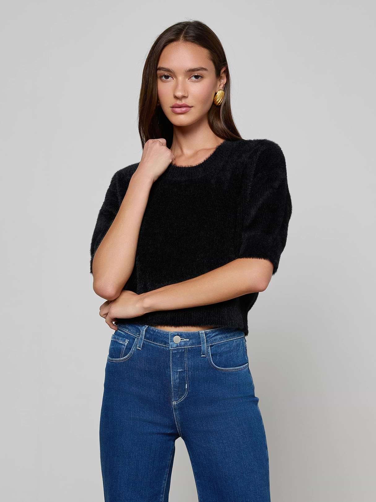 A woman with long brown hair stands confidently against a plain light background, wearing the ultra-soft Guinevere Cropped Pullover in black with short sleeves and blue jeans, one arm crossed while the other touches her face.