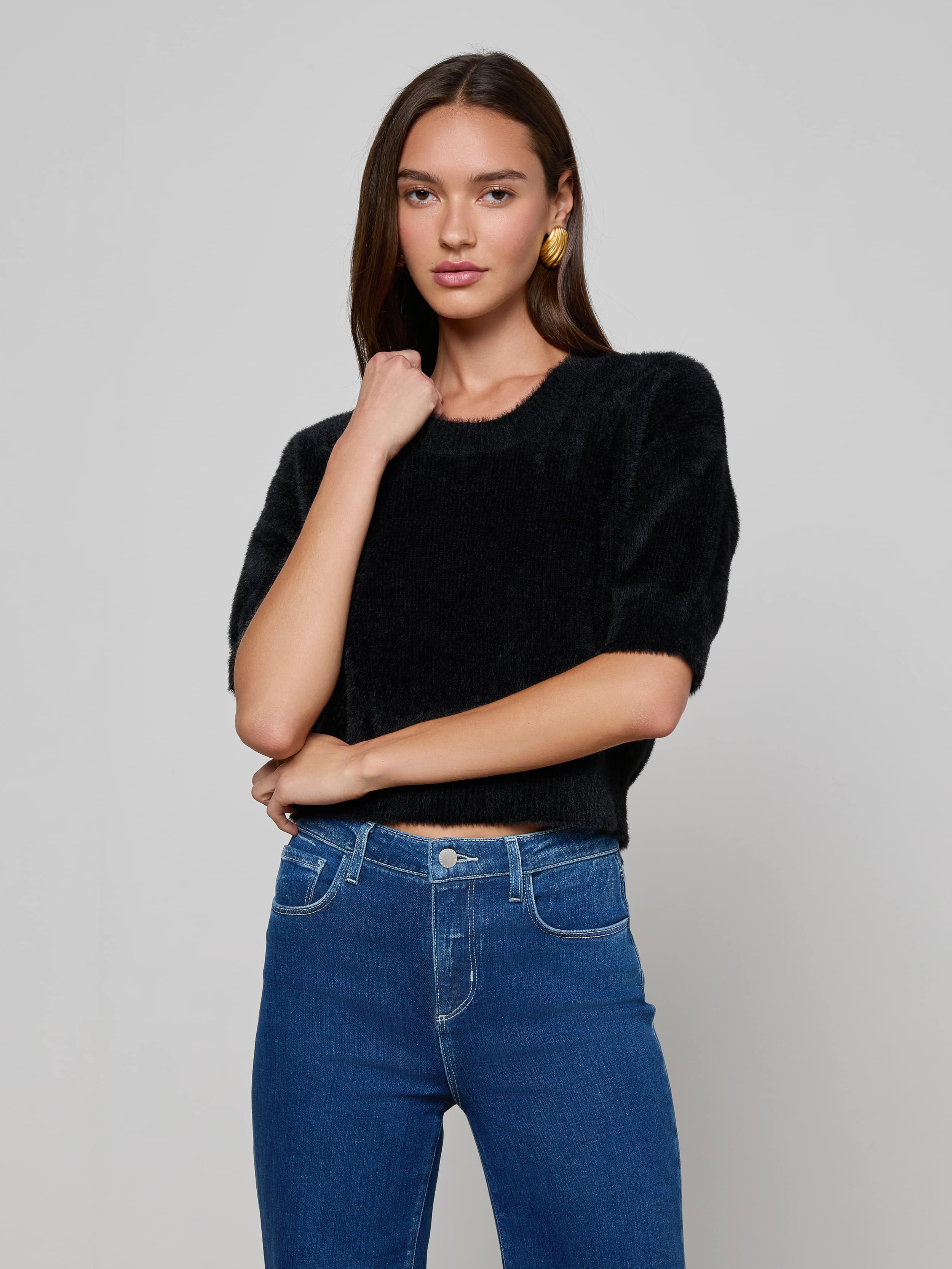 A woman with long brown hair stands confidently against a plain light background, wearing the ultra-soft Guinevere Cropped Pullover in black with short sleeves and blue jeans, one arm crossed while the other touches her face.