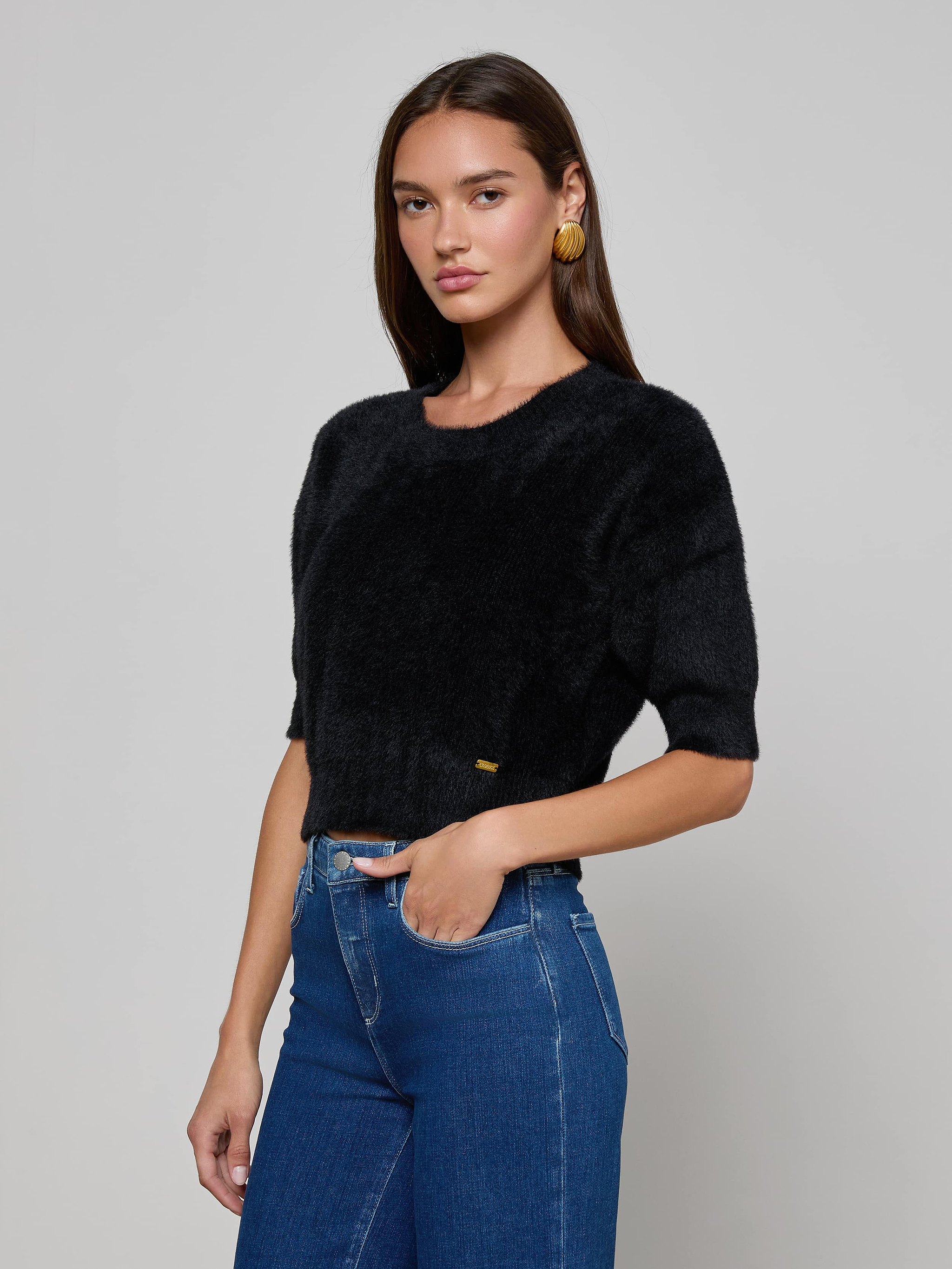 Wearing the Guinevere Cropped Pullover, a woman with long brown hair pairs the ultra-soft knit black short-sleeve crewneck sweater with blue high-waisted jeans and gold hoop earrings, standing against a plain light background.