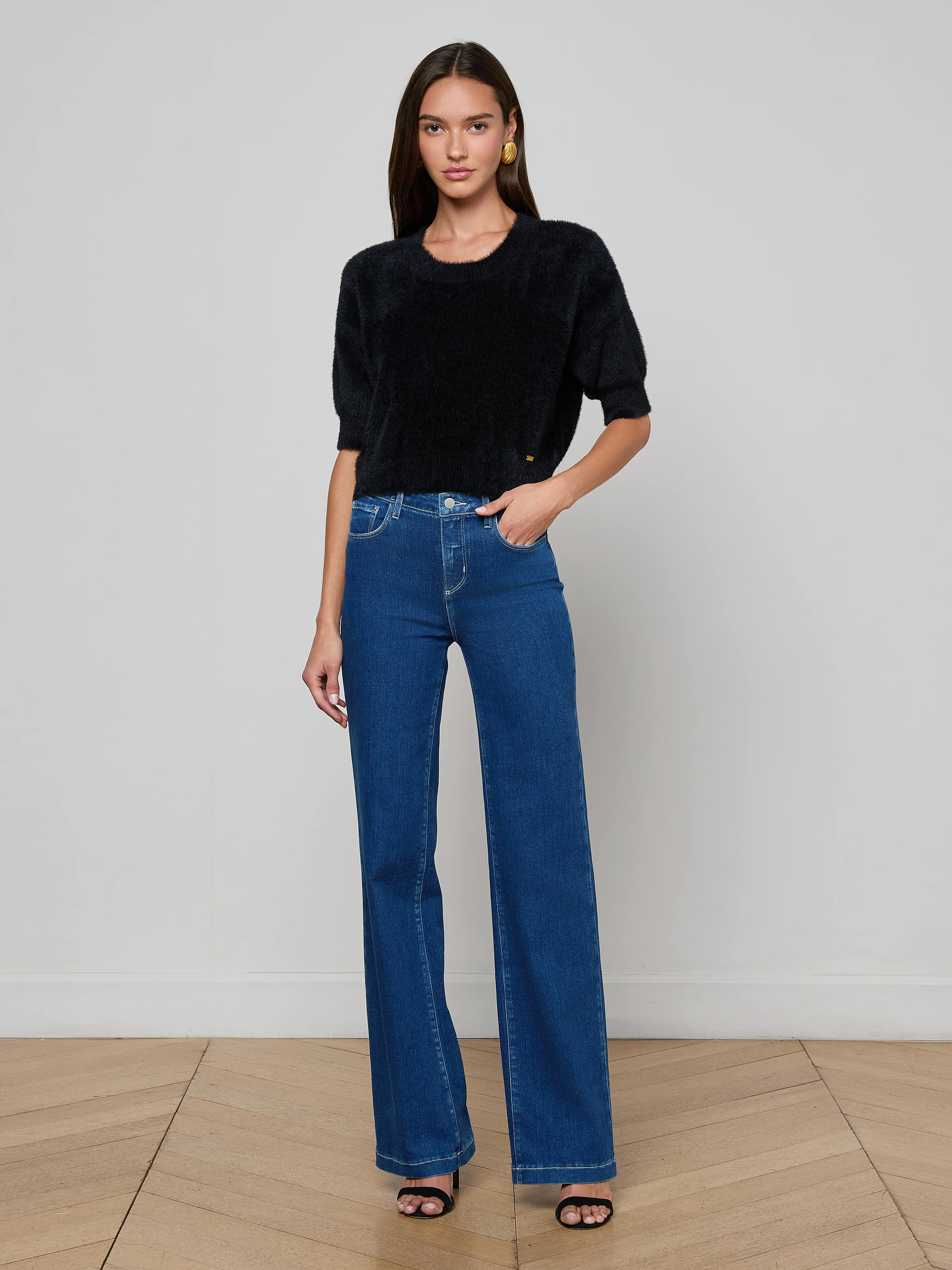 A woman stands facing forward, wearing the Guinevere Cropped Pullover in black paired with blue high-waisted wide-leg cropped jeans and black heeled sandals against a plain light background.