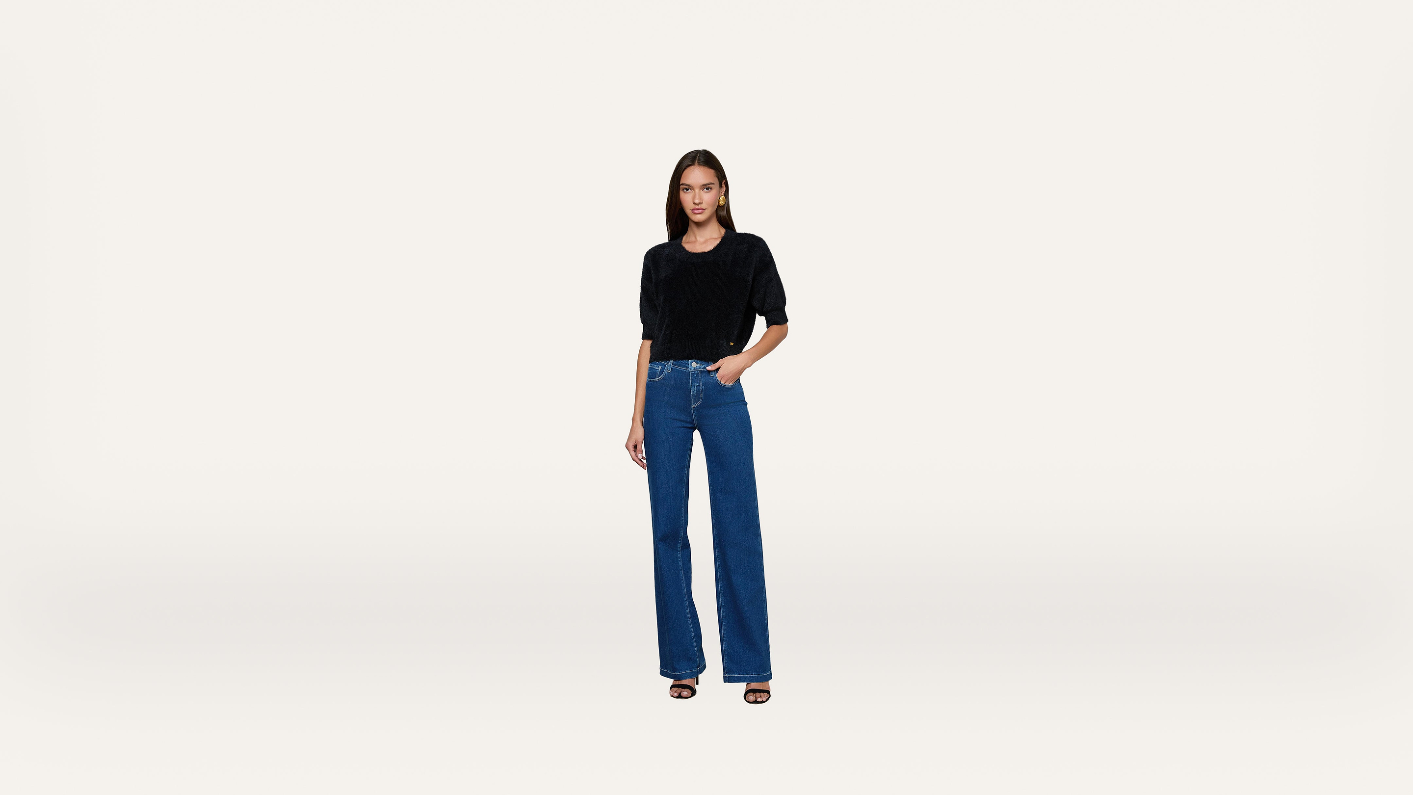 A woman stands facing forward, wearing a black top, blue high-waisted wide-leg jeans, and black heeled sandals, against a plain white background.