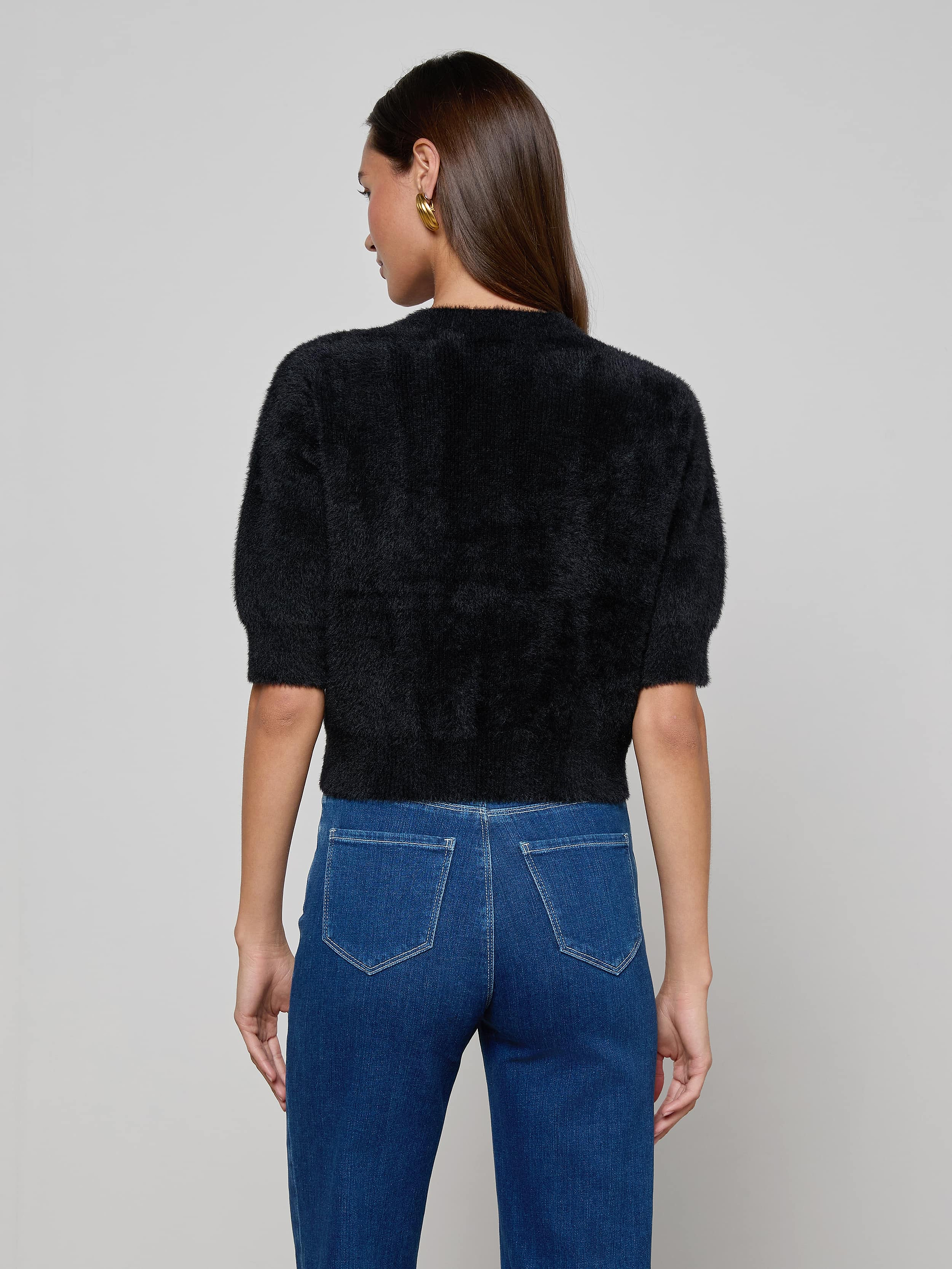 A person with long brown hair stands facing away, wearing the Guinevere Cropped Pullover—a fuzzy black crewneck sweater—paired with blue jeans against a plain light background.