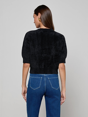 A woman with long brown hair wears the L’AGENCE Guinevere Cropped Pullover, a black fuzzy short-sleeve top, and blue jeans with back pockets. She faces away from the camera against a plain light gray background, accessorized with gold hoop earrings.
