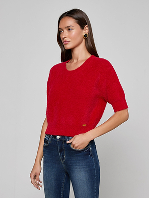 A woman with long brown hair models the Guinevere Cropped Pullover, an ultra-soft red knit crewneck sweater with short sleeves and a cropped fit, paired with blue jeans against a plain light gray background.