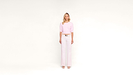 A woman stands against a plain white background, wearing a light pink top, white high-waisted wide-leg pants, and white sandals. She faces forward with a neutral expression.