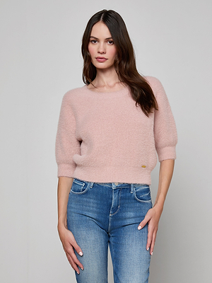 A woman stands against a plain background wearing the Guinevere Cropped Pullover, a light pink ultra-soft knit crewneck sweater with three-quarter sleeves, paired with blue jeans, looking at the camera with a neutral expression.