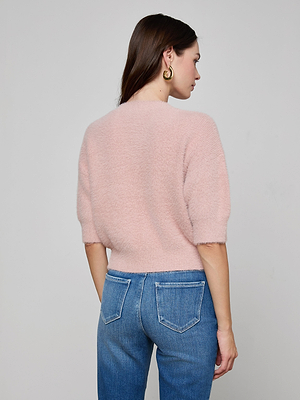A woman with long brown hair and gold hoop earrings wears the Guinevere Cropped Pullover—a soft knit, crewneck cropped sweater—paired with blue jeans, standing facing away from the camera against a plain gray background.