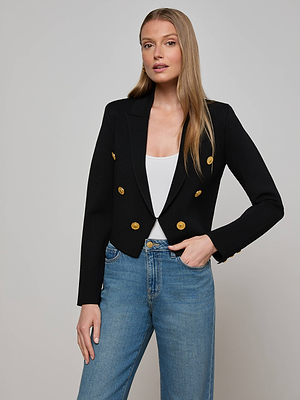 A woman with long, straight blonde hair wears the Elio Cropped Knit Blazer by L'AGENCE over a white top and blue jeans, standing against a light gray background with one hand in her pocket.
