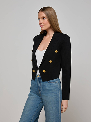A woman with straight, light brown hair models the L'AGENCE Elio Cropped Knit Blazer in black with gold buttons over a white top and blue jeans, standing against a plain light gray background.
