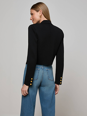 A woman with straight light brown hair wears the L'AGENCE Elio Cropped Knit Blazer, featuring gold sleeve buttons, paired with high-waisted blue jeans. She stands facing away against a plain light gray background.