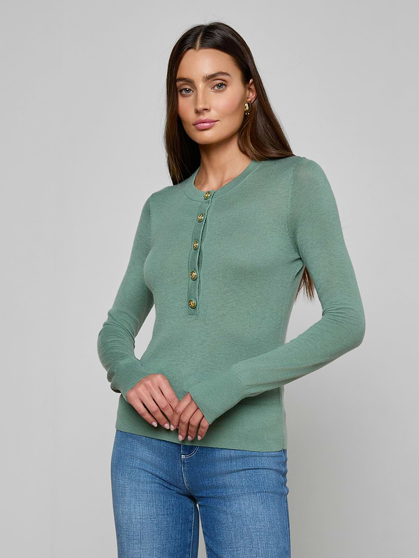 A woman with long brown hair wears the Ronda Knit Henley Top, a lightweight green classic sweater with a button placket, paired with blue jeans. She stands against a plain gray background, looking at the camera with a neutral expression.