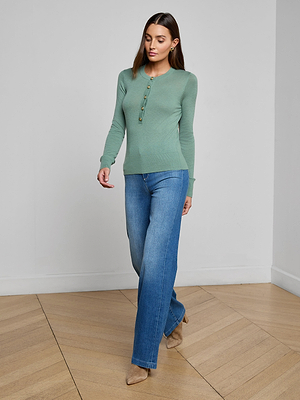A woman in the lightweight green Ronda Knit Henley Top and blue jeans walks indoors on a light wood floor, looking slightly downward with one hand by her side. She completes her look with tan pointed-toe shoes.
