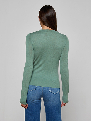 A person with long brown hair, wearing the Ronda Knit Henley Top in green and blue jeans, stands facing away from the camera against a plain light gray background.