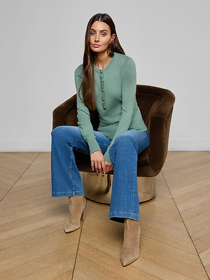 A woman with long brown hair sits on a brown velvet chair, wearing the Ronda Knit Henley Top in green, blue jeans, and tan heeled boots. The setting has light wood floors and a plain white wall.