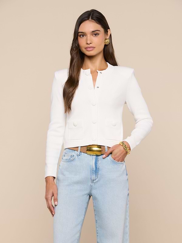 A woman with long brown hair wears the Patsy Cropped Cardigan—a white, buttoned style with subtle shoulder pads—paired with light blue jeans and gold accessories. She stands facing forward against a plain light background with a neutral expression.