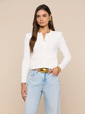 A woman with long brown hair stands against a beige background wearing the Patsy Cropped Cardigan, light blue jeans, gold bracelet and earrings, and a wide gold belt.