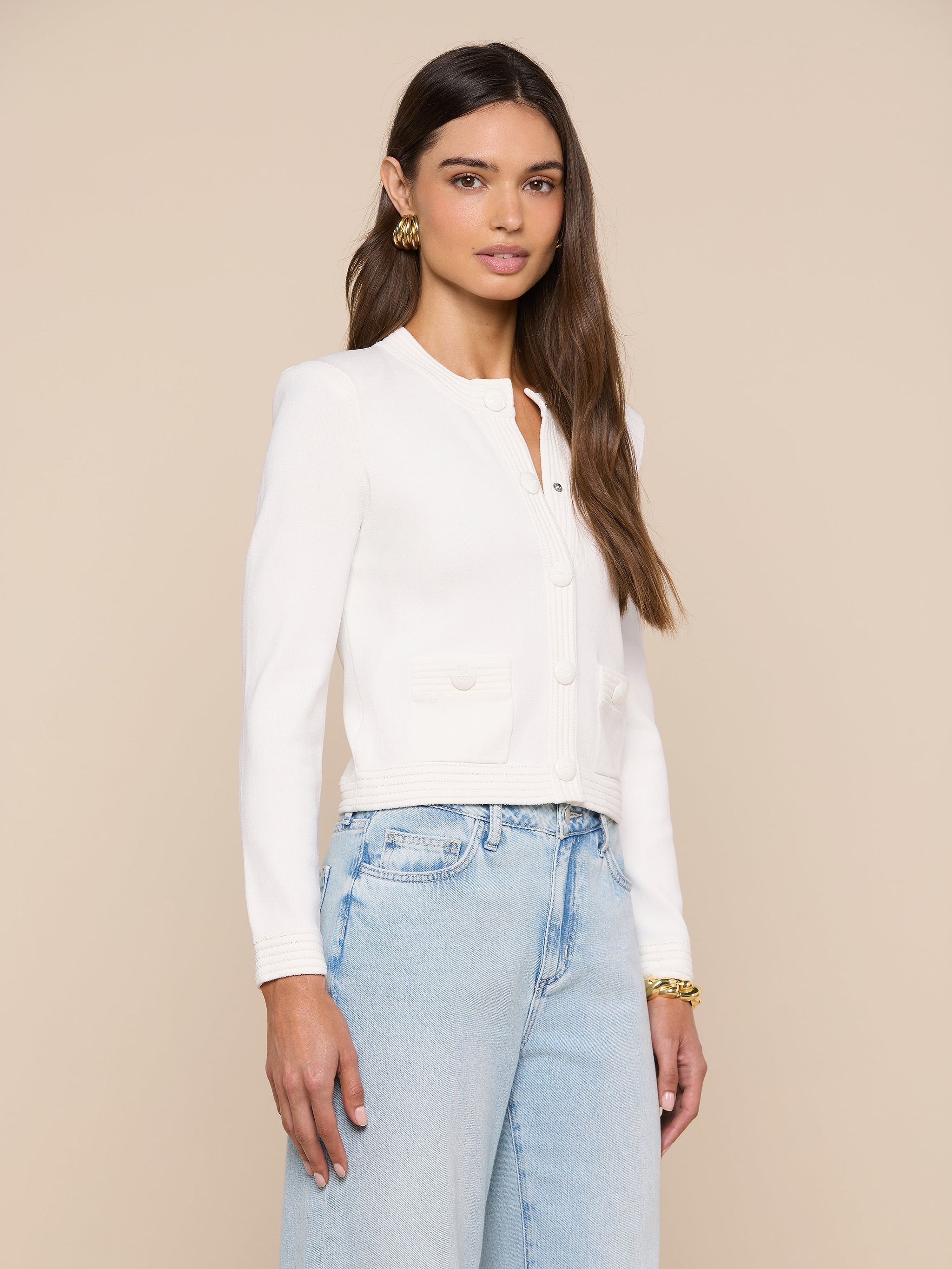 A woman with long brown hair stands confidently against an off-white background, wearing the Patsy Cropped Cardigan—a white jacket featuring ribbed trims—layered over a white top and paired with light blue high-waisted jeans.