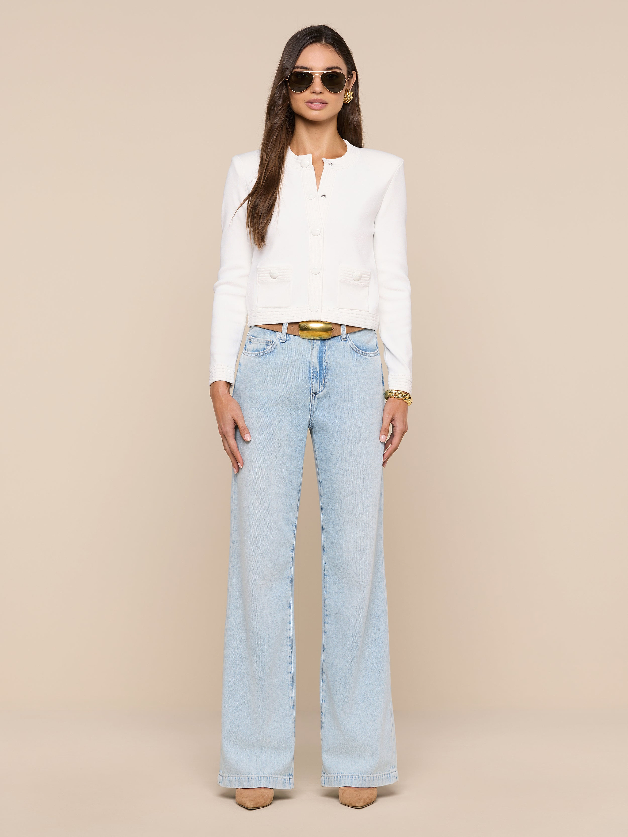 Woman in the Patsy Cropped Cardigan—a white long-sleeve top with ribbed trims—paired with light blue wide-leg jeans, tan heels, and sunglasses stands against a plain white background. She has long hair down and accessorizes with a belt and bracelets.