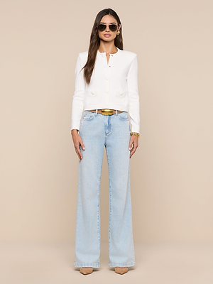 A woman wearing sunglasses, the Patsy Cropped Cardigan over an ivory top with ribbed trims, light blue high-waisted wide-leg jeans, beige heels, a brown belt, and a gold bracelet stands against a beige background.