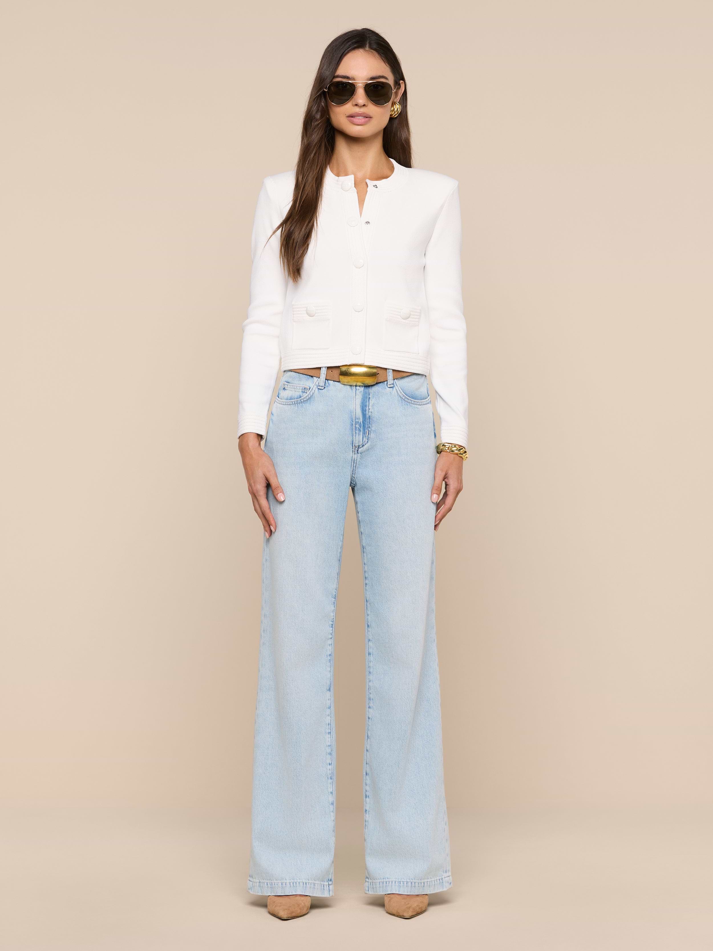 Woman in the Patsy Cropped Cardigan—a white long-sleeve top with ribbed trims—paired with light blue wide-leg jeans, tan heels, and sunglasses stands against a plain white background. She has long hair down and accessorizes with a belt and bracelets.