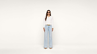 A woman stands against a plain white background wearing sunglasses, a white long-sleeve top, light blue high-waisted wide-leg jeans, and beige heels.