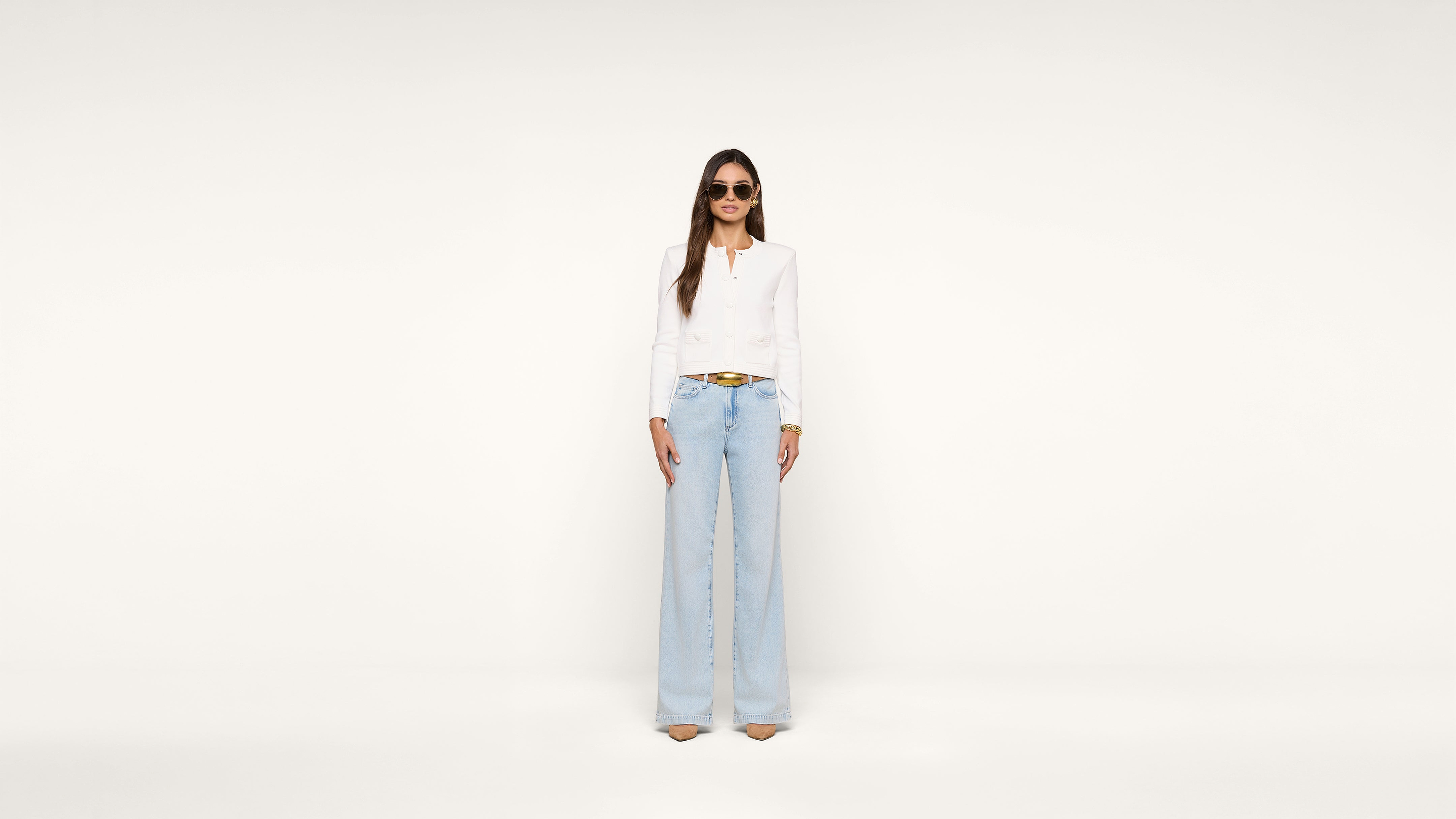 A woman stands against a plain white background wearing sunglasses, a white long-sleeve top, light blue high-waisted wide-leg jeans, and beige heels.