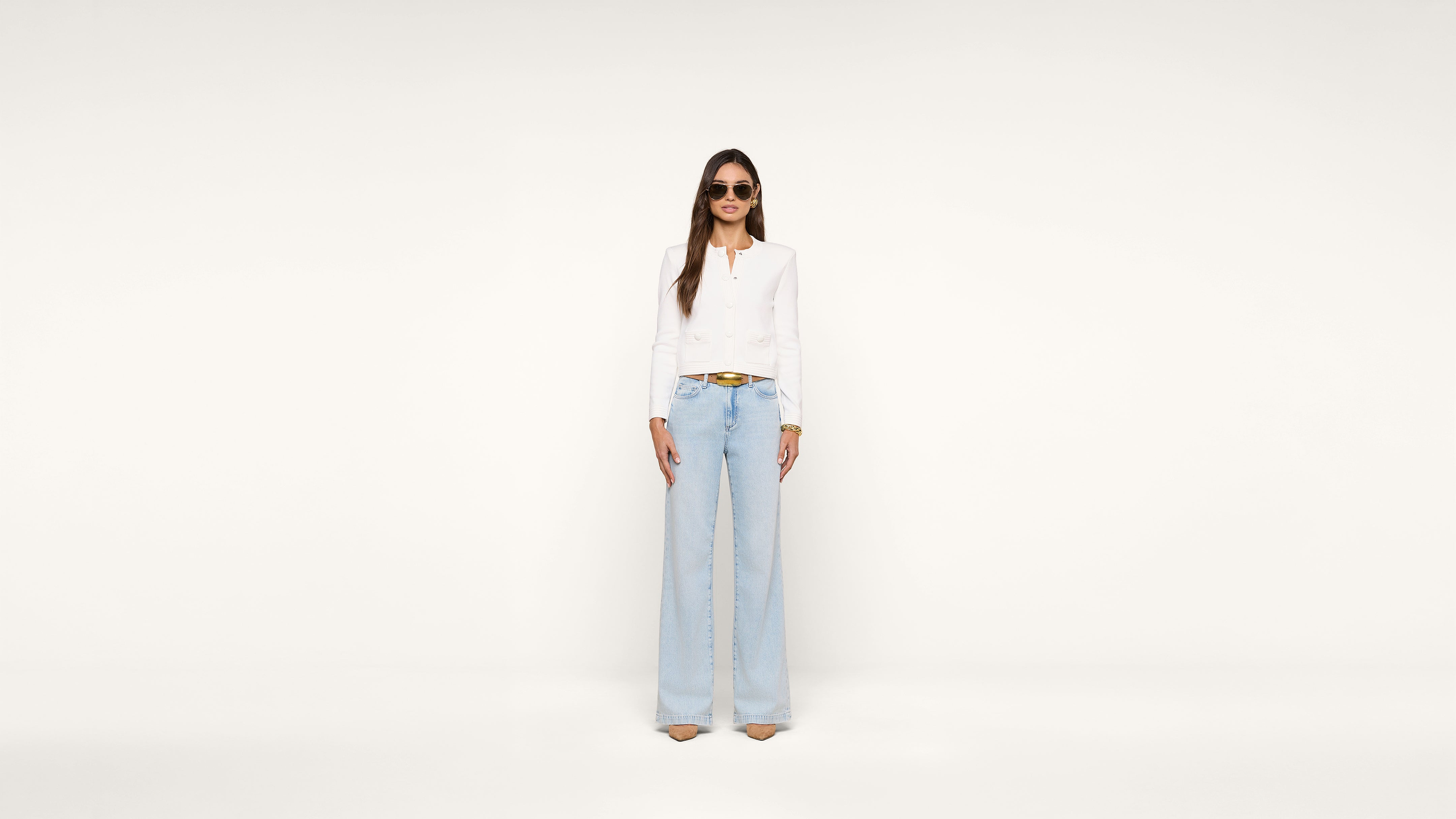 A woman stands against a plain white background wearing sunglasses, a white long-sleeve top, light blue high-waisted wide-leg jeans, and beige heels.