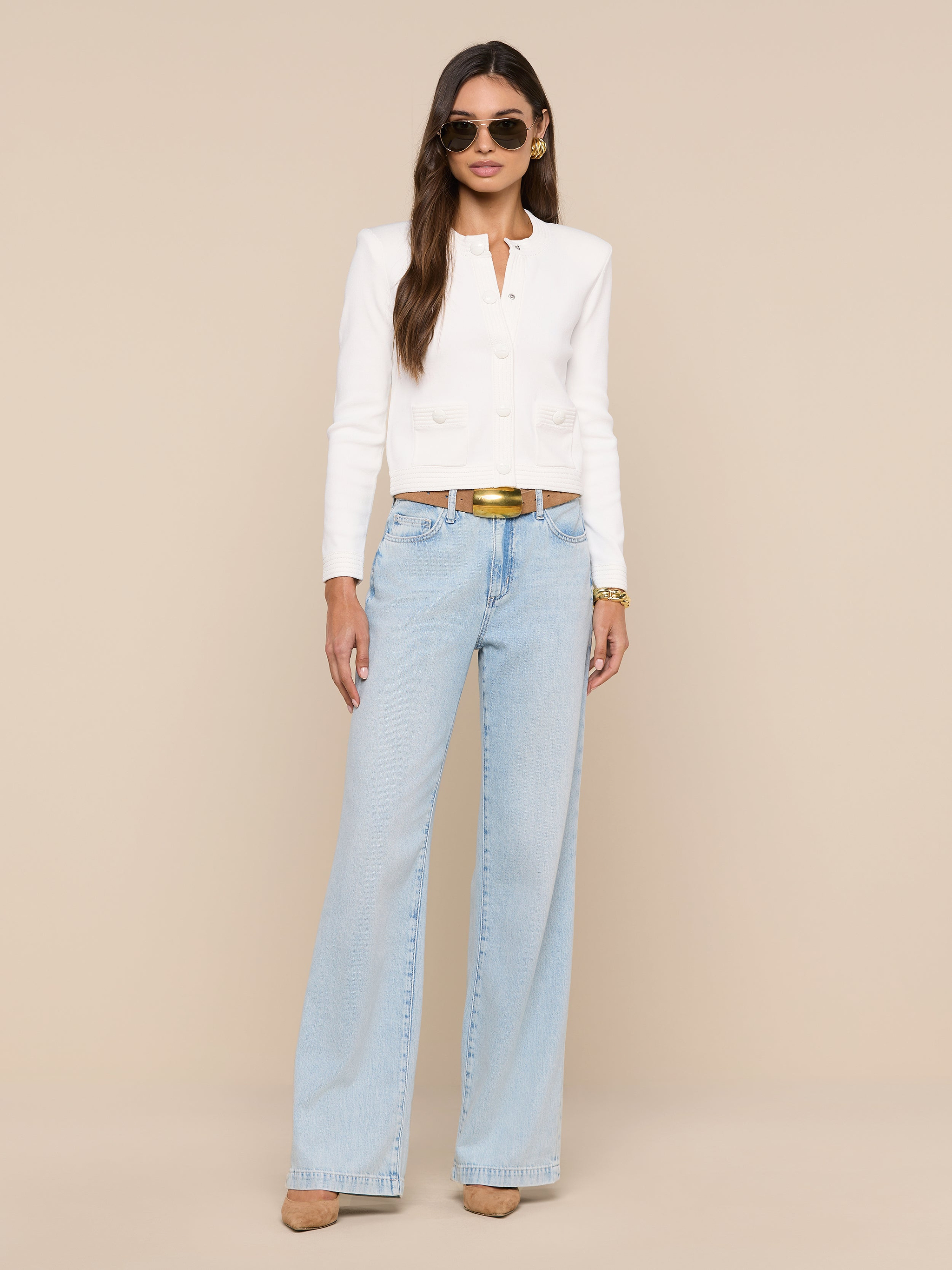 A woman stands against a white background wearing sunglasses, the Patsy Cropped Cardigan with ribbed trims, light blue wide-leg jeans, a brown belt, and beige heels. She has long, straight hair and her hands are by her sides.