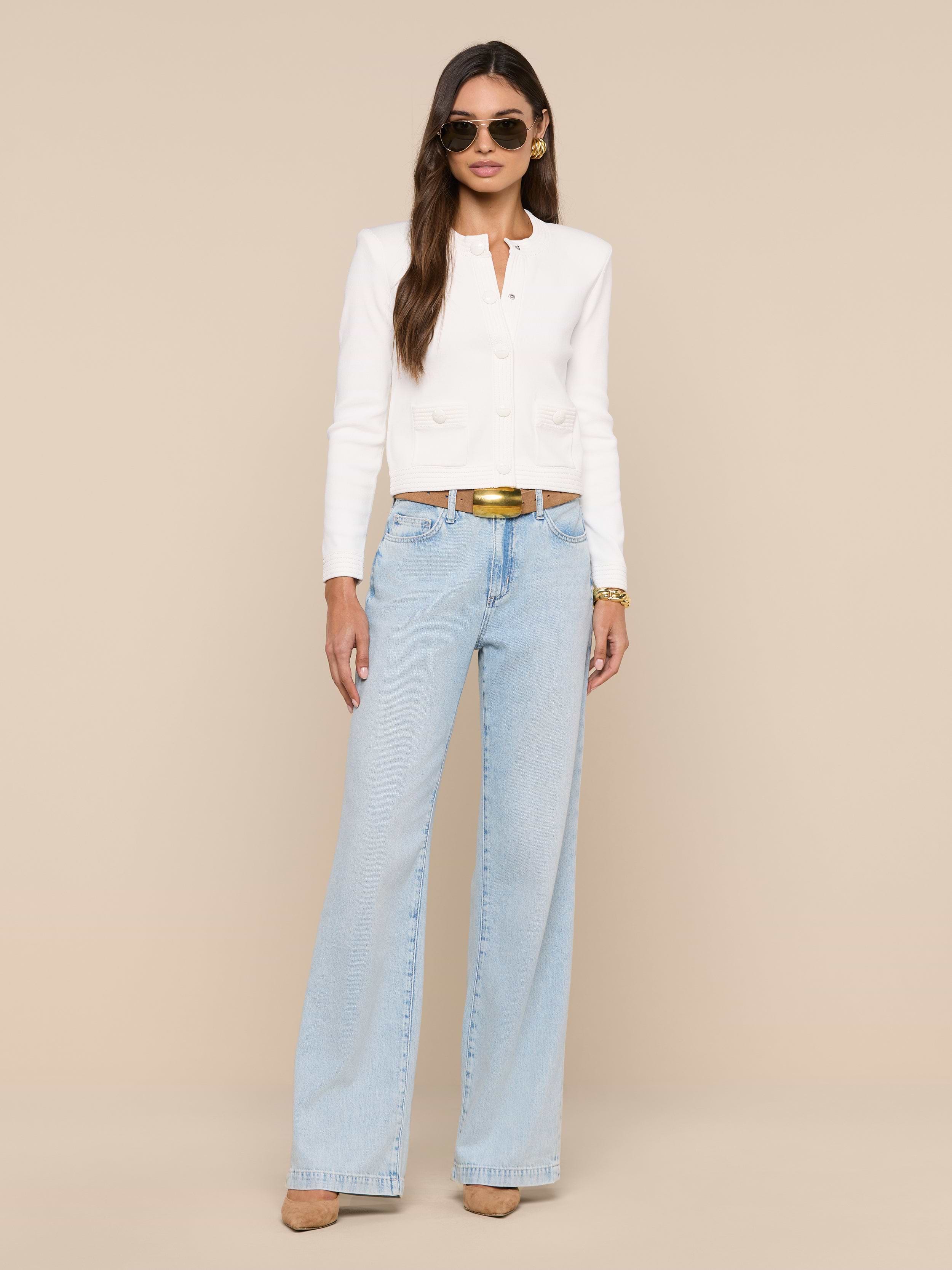 A woman stands against a white background wearing sunglasses, the Patsy Cropped Cardigan with ribbed trims, light blue wide-leg jeans, a brown belt, and beige heels. She has long, straight hair and her hands are by her sides.