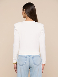 A person with long brown hair is shown from behind, wearing the Patsy Cropped Cardigan—a white long-sleeve top with ribbed trims—paired with light blue jeans. Gold earrings and a bracelet are visible. The background is plain and light-colored.