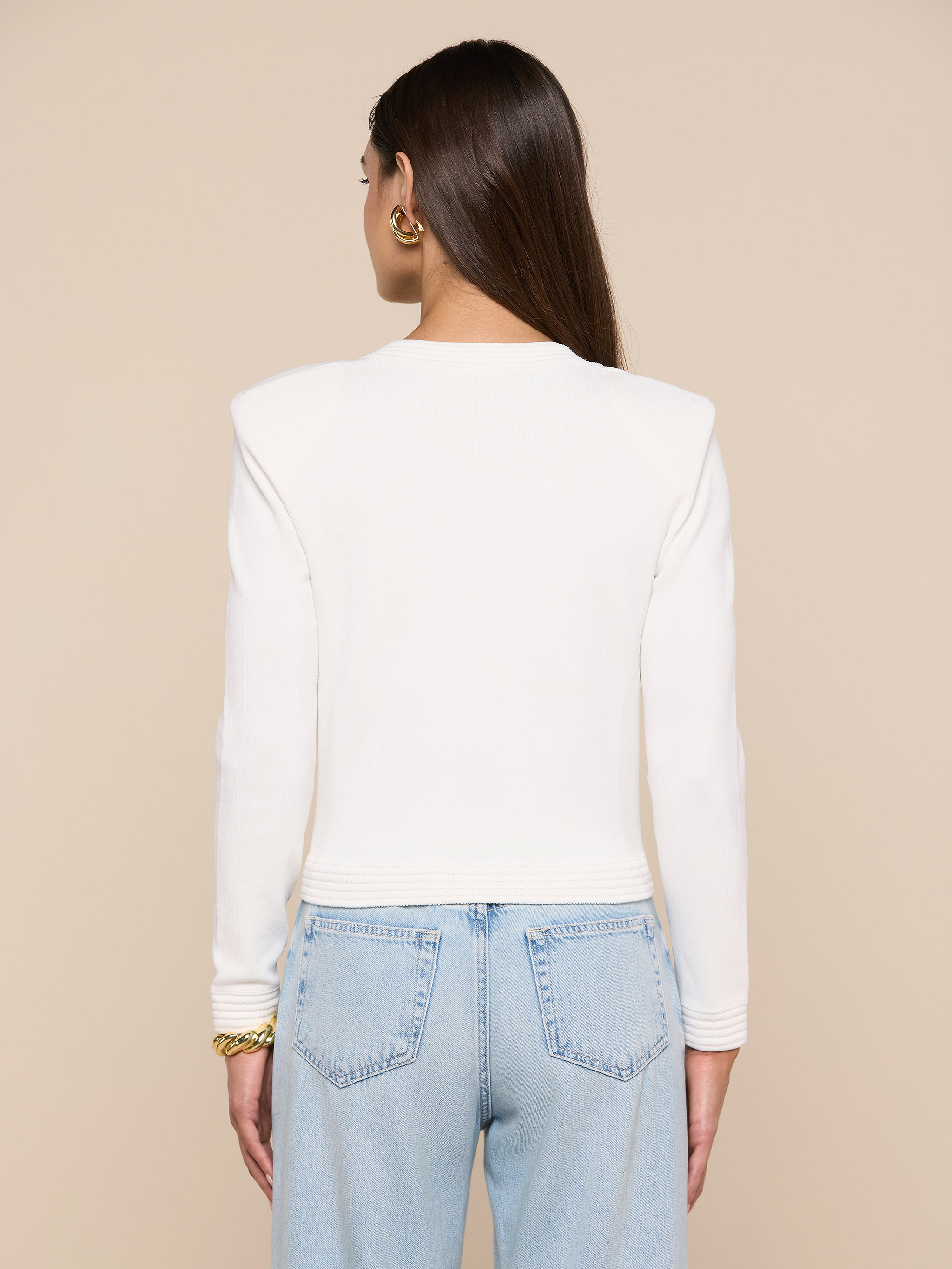 A person with long brown hair is shown from behind, wearing the Patsy Cropped Cardigan—a white long-sleeve top with ribbed trims—paired with light blue jeans. Gold earrings and a bracelet are visible. The background is plain and light-colored.
