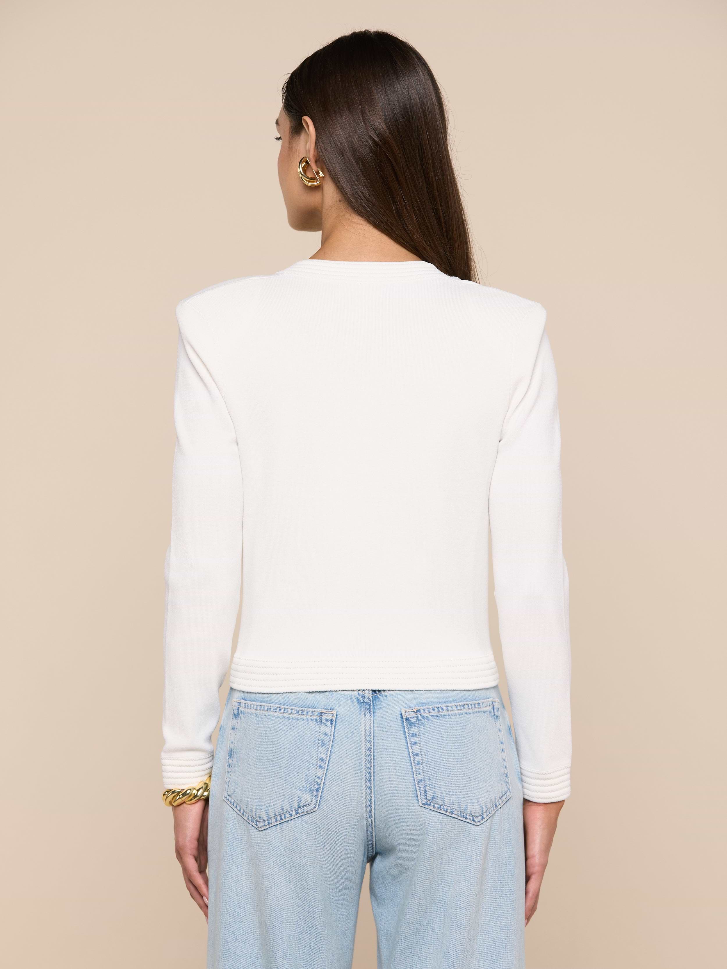 A person with long brown hair is shown from behind, wearing the Patsy Cropped Cardigan—a white long-sleeve top with ribbed trims—paired with light blue jeans. Gold earrings and a bracelet are visible. The background is plain and light-colored.