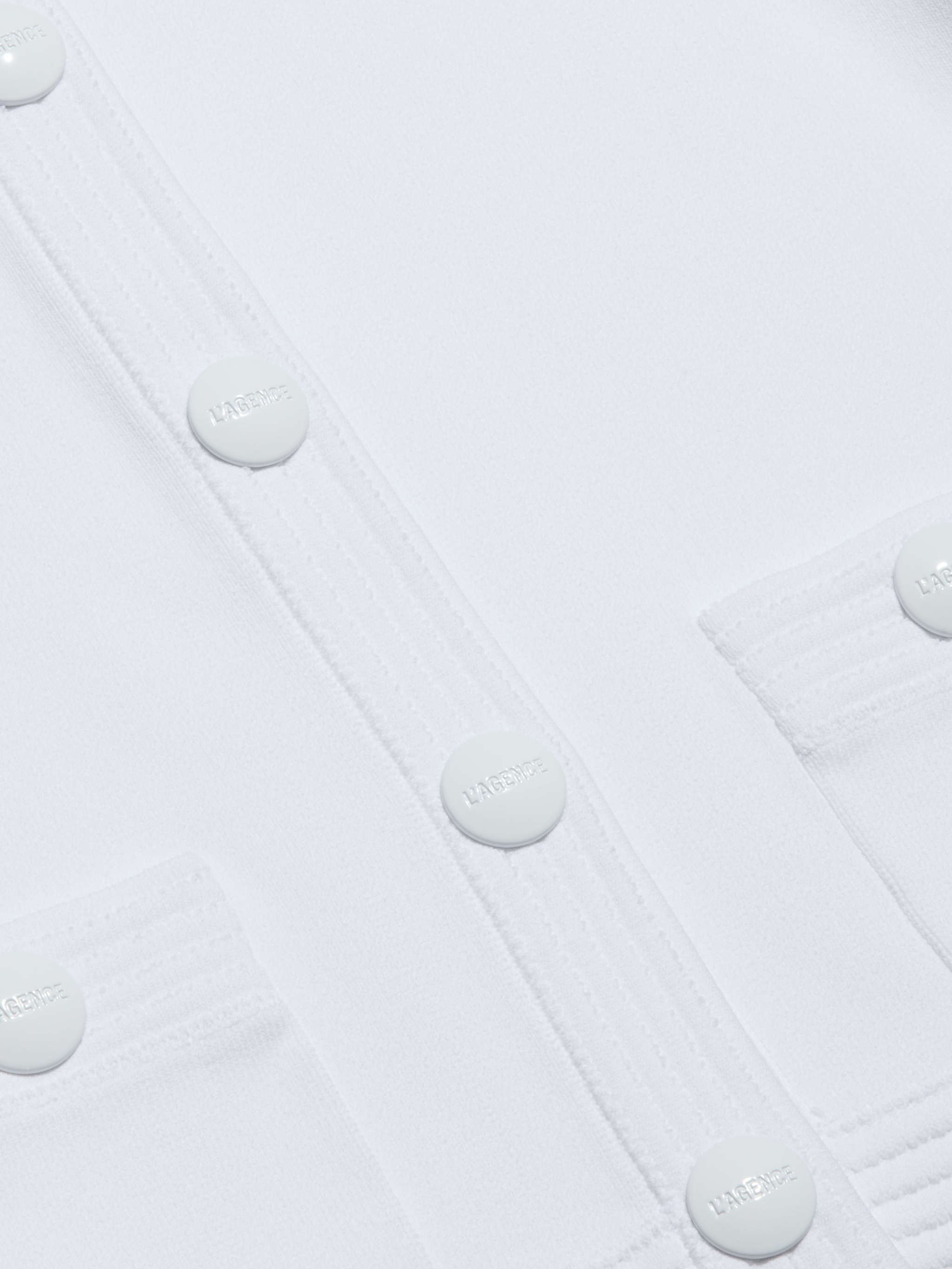 Close-up of the Patsy Cropped Cardigan in white, featuring round buttons embossed with the word HARMONIE and stitched pocket flaps. The smooth fabric showcases visible seam lines, while subtle shoulder pads add structure to the design.
