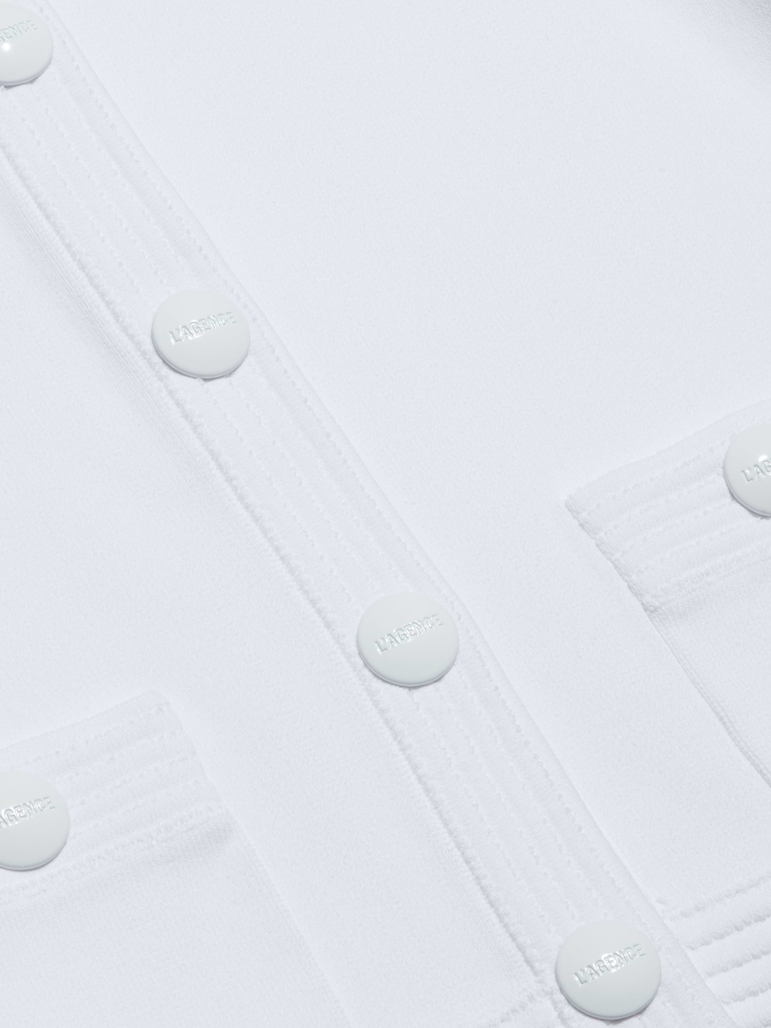 Close-up of the Patsy Cropped Cardigan in white, featuring round buttons embossed with the word HARMONIE and stitched pocket flaps. The smooth fabric showcases visible seam lines, while subtle shoulder pads add structure to the design.