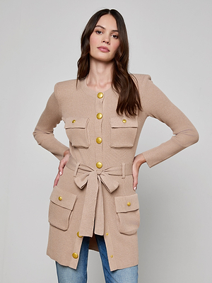 A woman with long brown hair and a neutral expression stands with her hands on her hips, wearing the beige Lore Cardigan featuring gold buttons, four front pockets, a tie belt at the waist, and blue jeans.