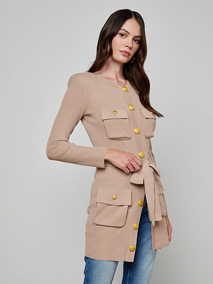 A woman with long brown hair models the Lore Cardigan, a beige knit sweater featuring large gold buttons and front pockets, paired with blue jeans against a plain light background.