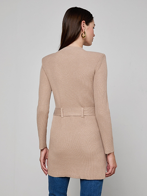 A woman with straight brown hair and gold hoop earrings stands against a plain light background, wearing the beige Lore Cardigan with a tie belt and blue jeans.