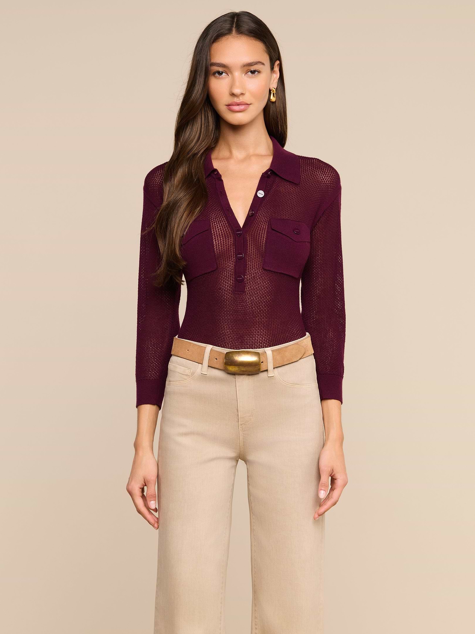 Una donna è in piedi, rivolta verso la fotocamera, e indossa un top Carine color bordeaux con bottoni e tasche funzionali, pantaloni beige a vita alta e una cintura color cuoio con fibbia dorata, su uno sfondo beige a tinta unita.