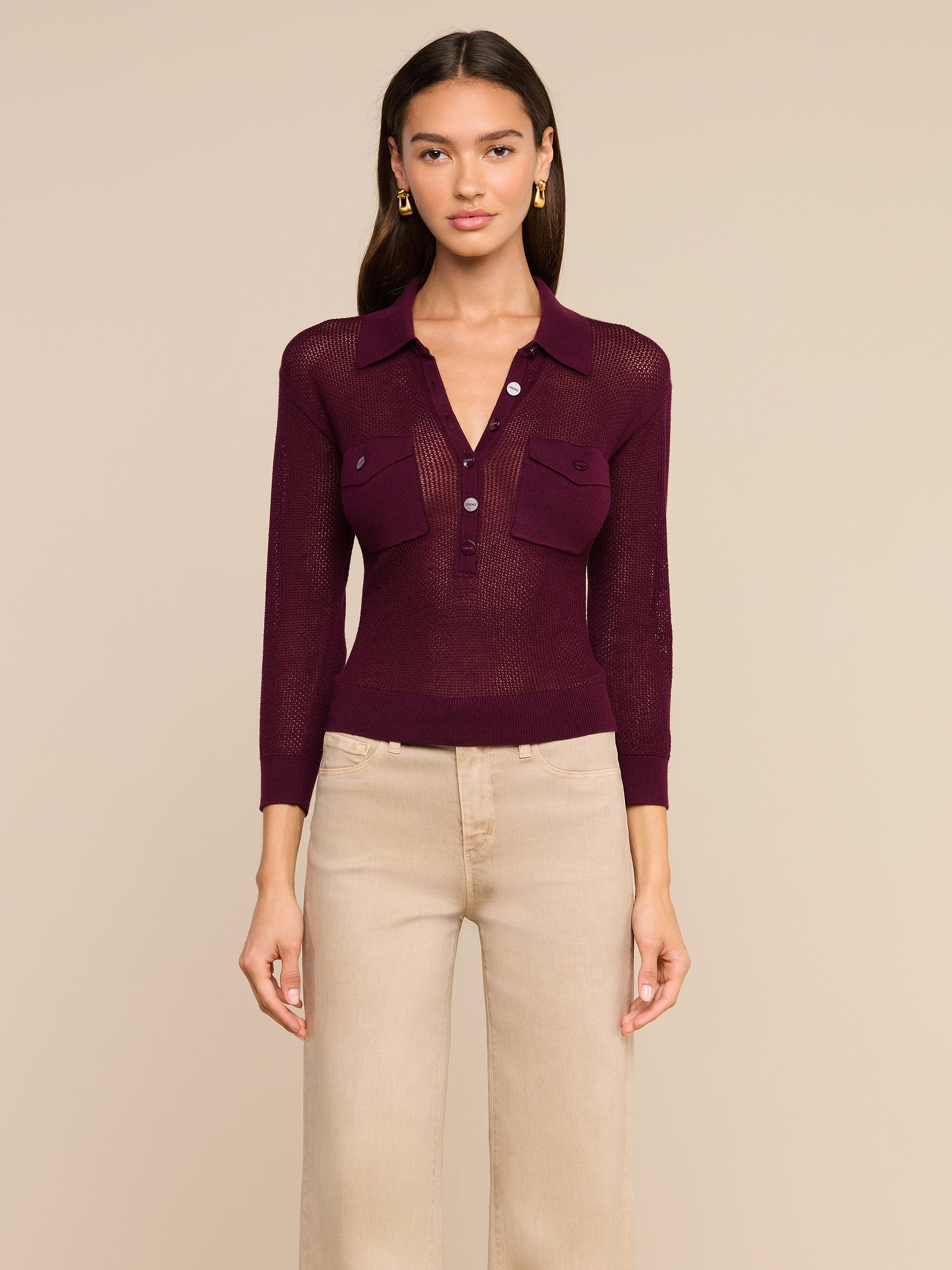 A woman stands facing forward wearing the Carine Top, a sheer burgundy collared blouse with utility pockets and buttons, paired with high-waisted beige pants. She has long dark hair and gold earrings. The background is plain and light-colored.