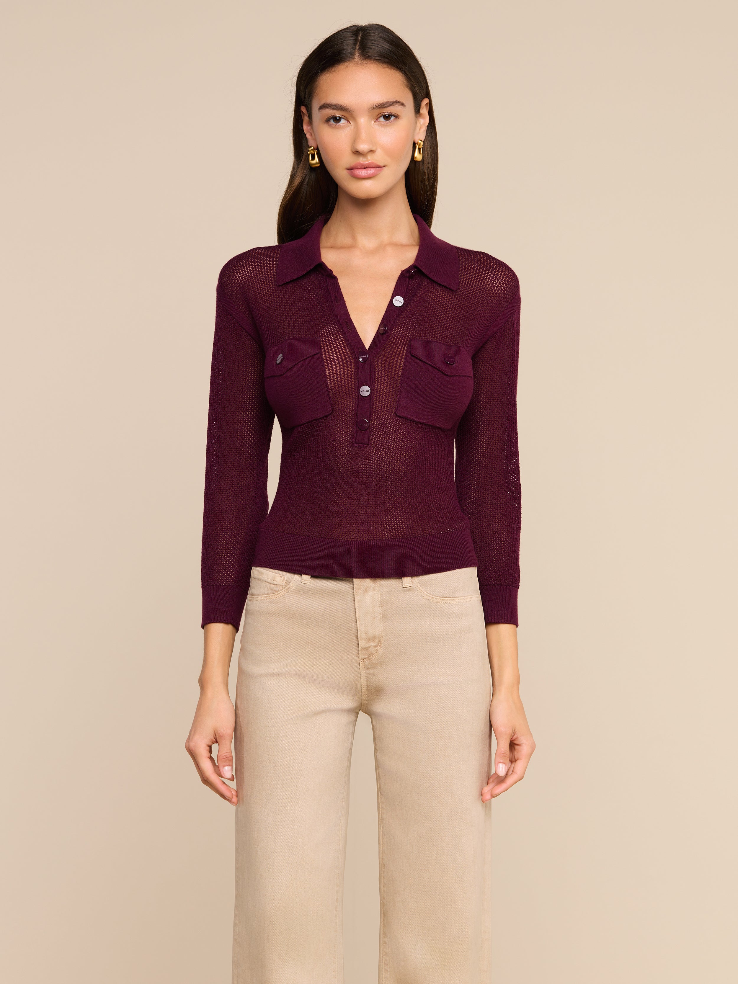 A woman stands facing forward wearing the Carine Top, a sheer burgundy collared blouse with utility pockets and buttons, paired with high-waisted beige pants. She has long dark hair and gold earrings. The background is plain and light-colored.