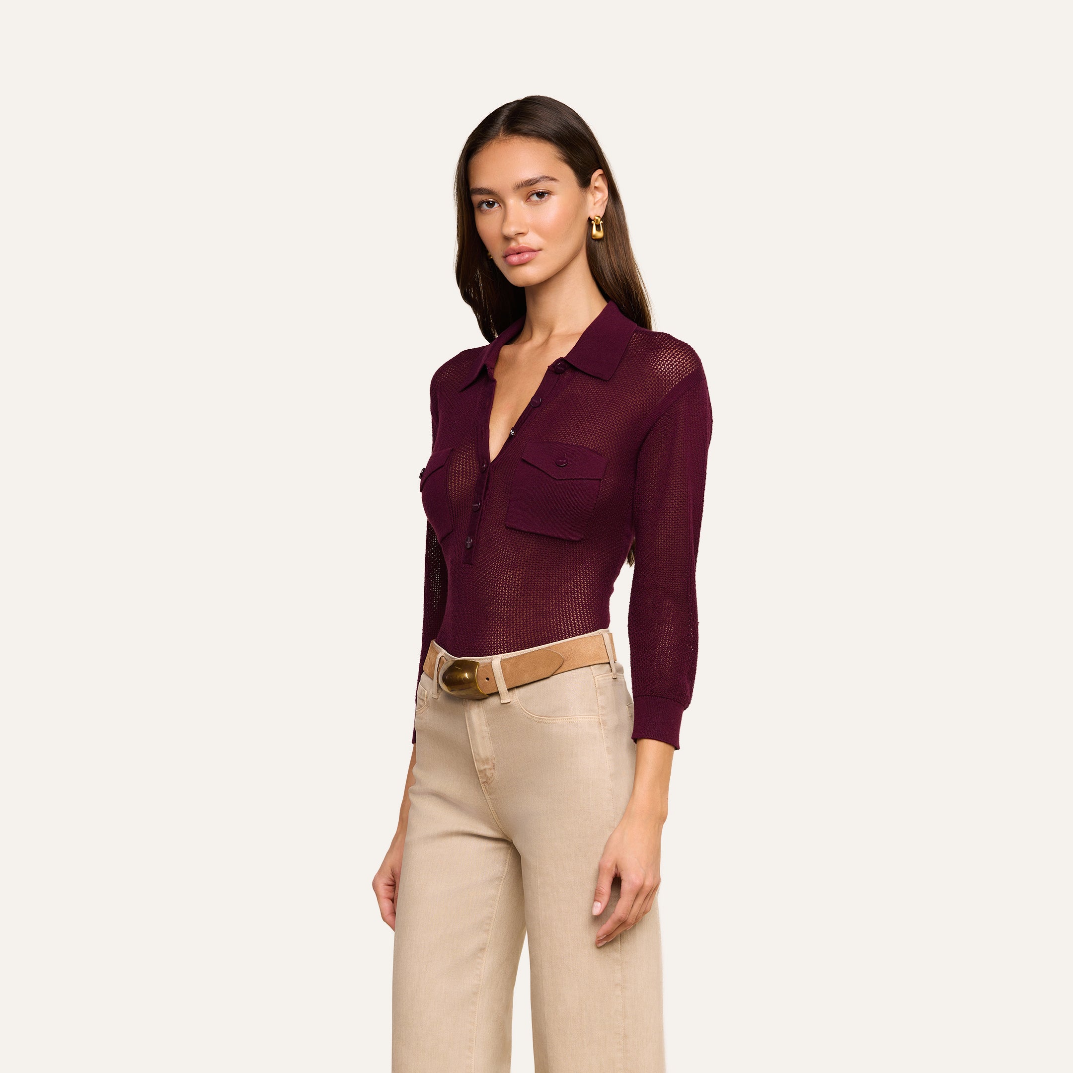 Wearing the Carine Top, a sheer burgundy button-up with utility pockets, paired with beige high-waisted pants, a tan belt, and gold earrings, she stands against a plain background.