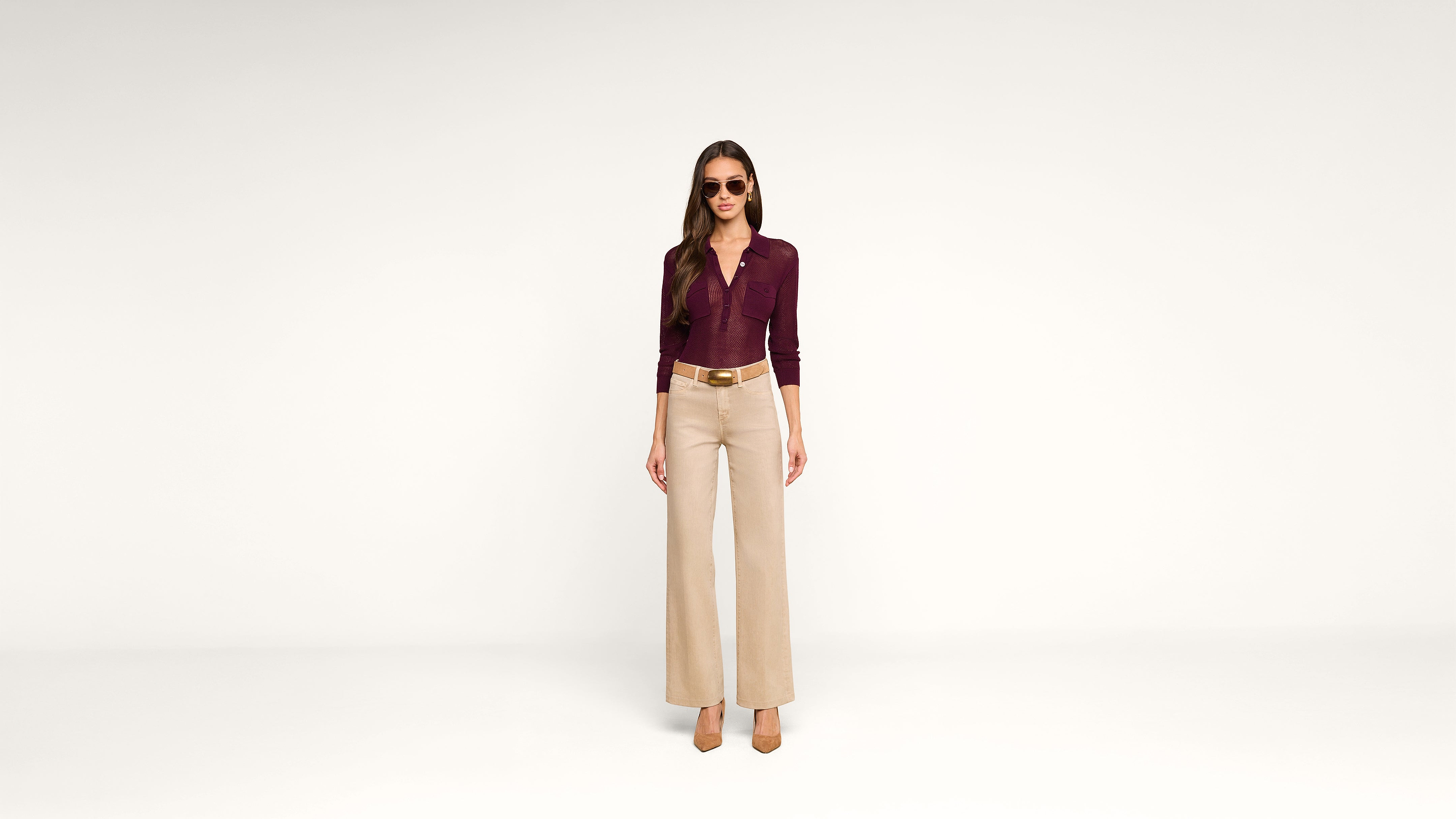 A woman wearing sunglasses, a maroon button-up top, beige high-waisted wide-leg pants, and brown heels stands against a plain white background.