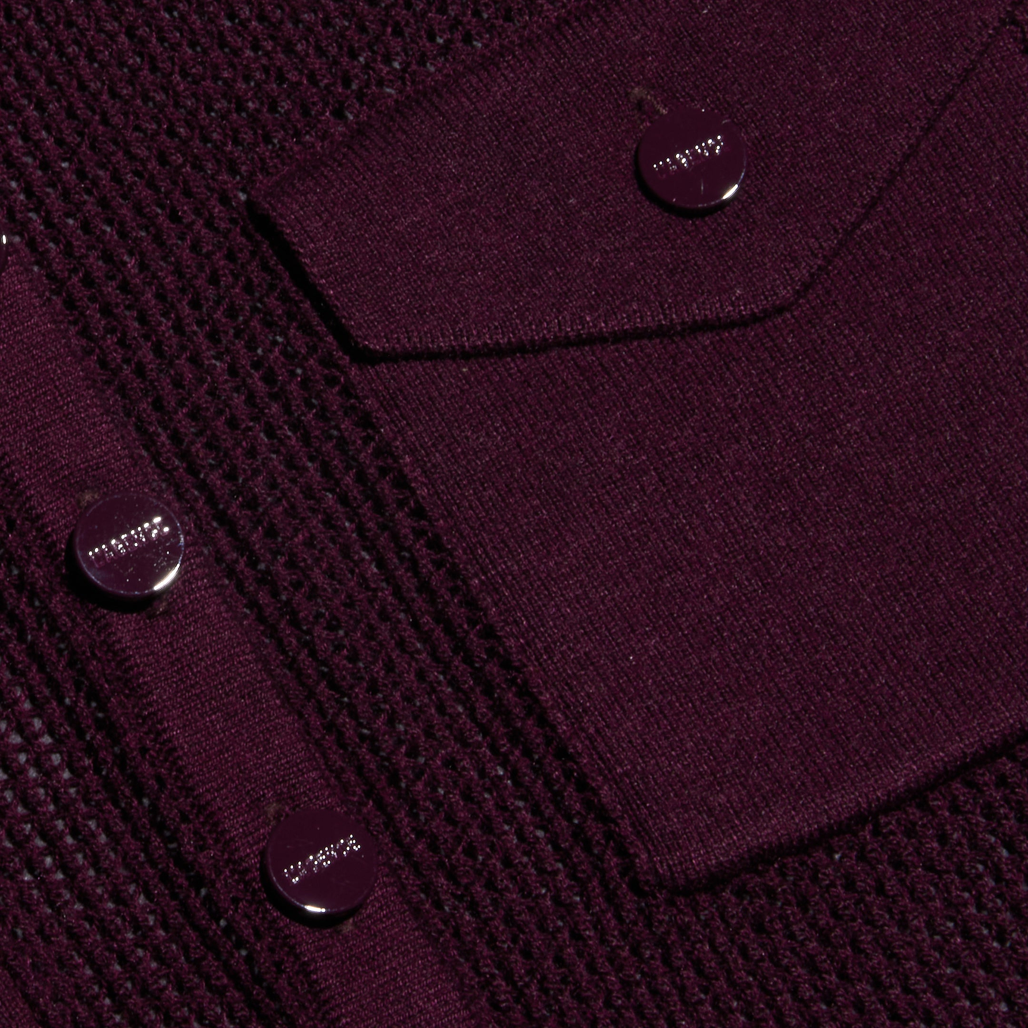 Close-up of a textured, dark purple fabric featuring a utility pocket with a flap and shiny round buttons. The lightweight knit fabric has a distinctive pattern along with smoother sections, reminiscent of the Carine Top.
